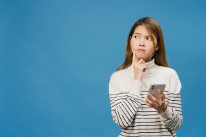 thinking-dreaming-young-asia-lady-using-phone-with-positive-expression-dressed-casual-clothing-feeling-happiness-stand-isolated-blue-wall
